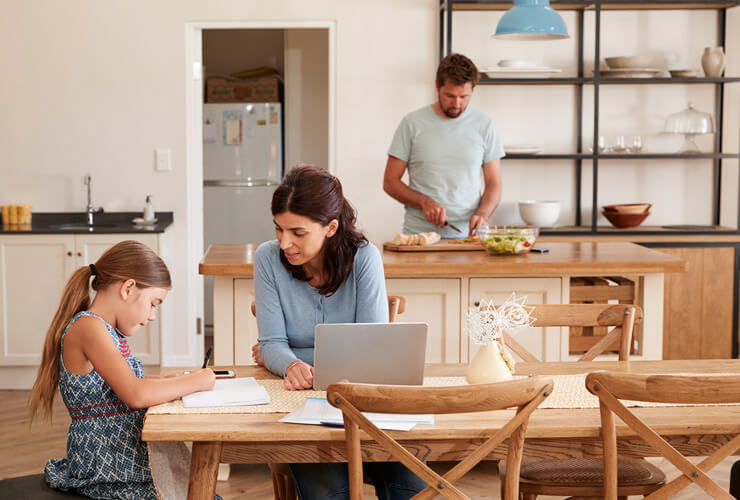 Mom on laptop helps daughter with homework at kitchen table while Dad fixes dinner